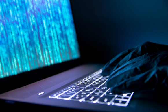 Imagen de manos de un ciberdelincuente con guantes negros digitando un teclado de un portátil encendido.