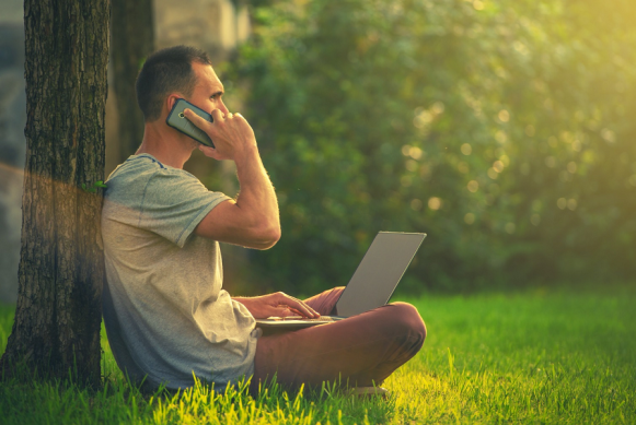 Imagen de un hombre en un parque sentado sobre la grama manipulando su computadora y hablando por su móvil.