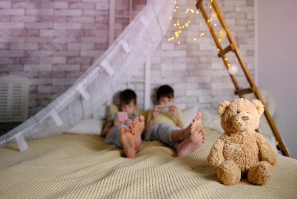 Fotografía de dos niños acostados en una cama manipulando sus dispositivos móviles e ignorando un oso de peluche.